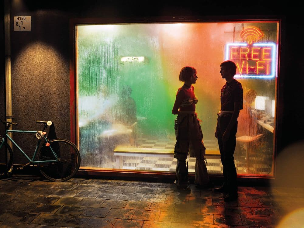 Two silhouetted figures on a night street outside a condensation-covered café window, neon light glowing within.